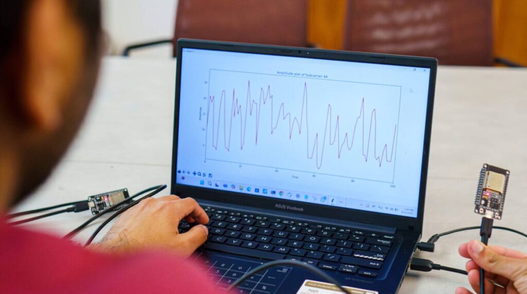 Researcher viewing heart rate data on laptop screen with WiFi hardware devices connected, showing the Pulse-Fi system monitoring heart rate without wearable devices.