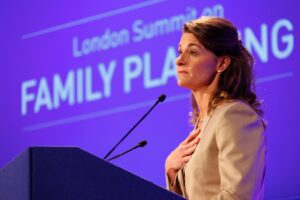 Melinda French Gates stands at a podium, speaking with her hand over her heart. Behind her, a purple screen displays the words 'London Summit on Family Planning'.