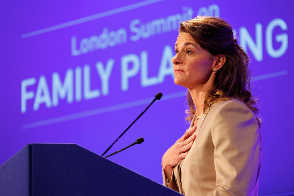 Melinda French Gates stands at a podium, speaking with her hand over her heart. Behind her, a purple screen displays the words 'London Summit on Family Planning'.