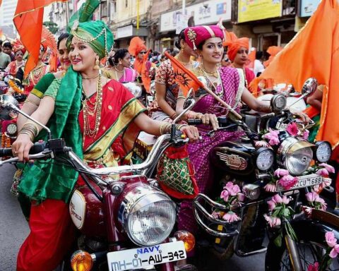 Women wearing colorful traditional Maharashtrian sarees and ornate jewelry riding decorated motorcycles with flowers during Gudi Padwa festival procession in Maharashtra, India.