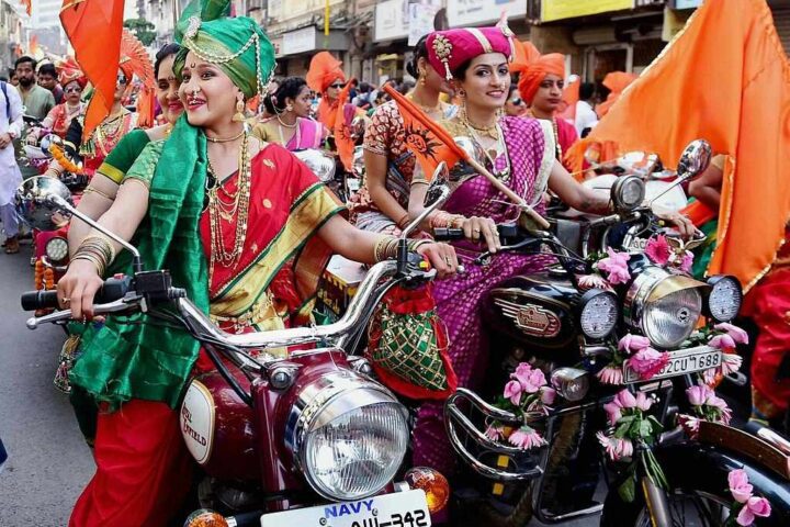 Women wearing colorful traditional Maharashtrian sarees and ornate jewelry riding decorated motorcycles with flowers during Gudi Padwa festival procession in Maharashtra, India.