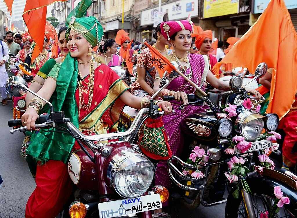 Women wearing colorful traditional Maharashtrian sarees and ornate jewelry riding decorated motorcycles with flowers during Gudi Padwa festival procession in Maharashtra, India.