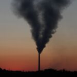 Dark plume of smoke billowing from a tall industrial chimney stack at the Łaziska thermal power plant silhouetted against an orange sunset sky.