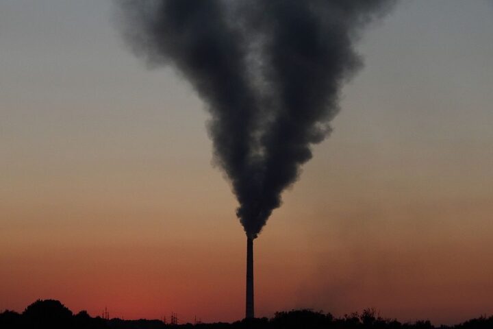 Dark plume of smoke billowing from a tall industrial chimney stack at the Łaziska thermal power plant silhouetted against an orange sunset sky.