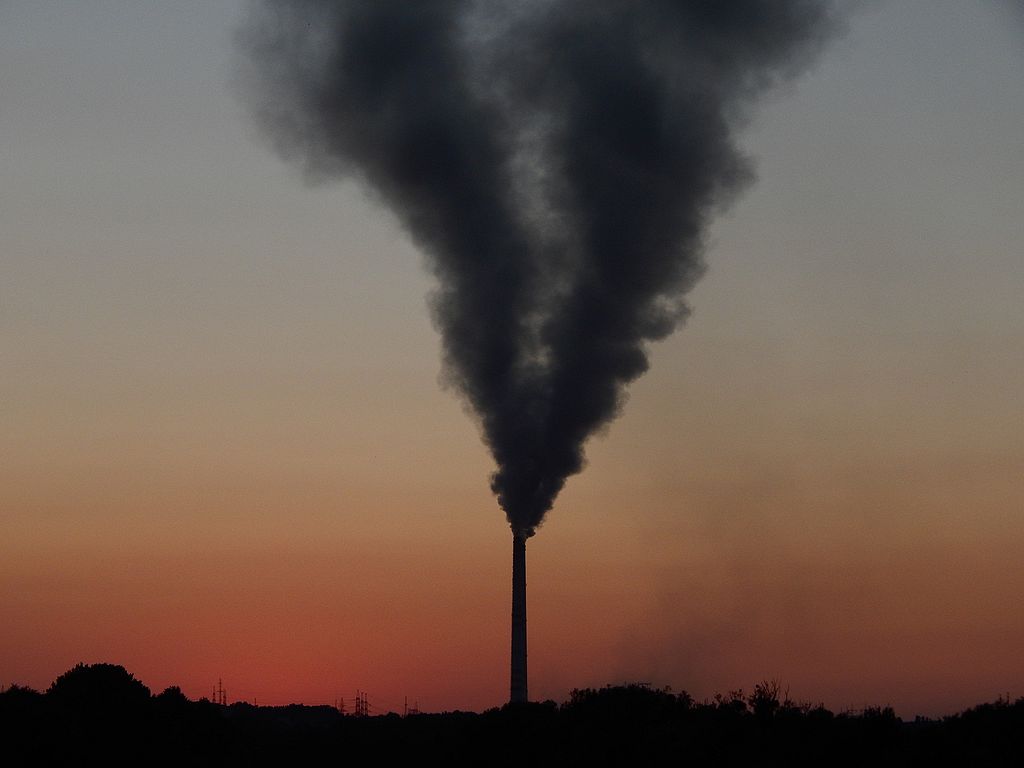 Dark plume of smoke billowing from a tall industrial chimney stack at the Łaziska thermal power plant silhouetted against an orange sunset sky.