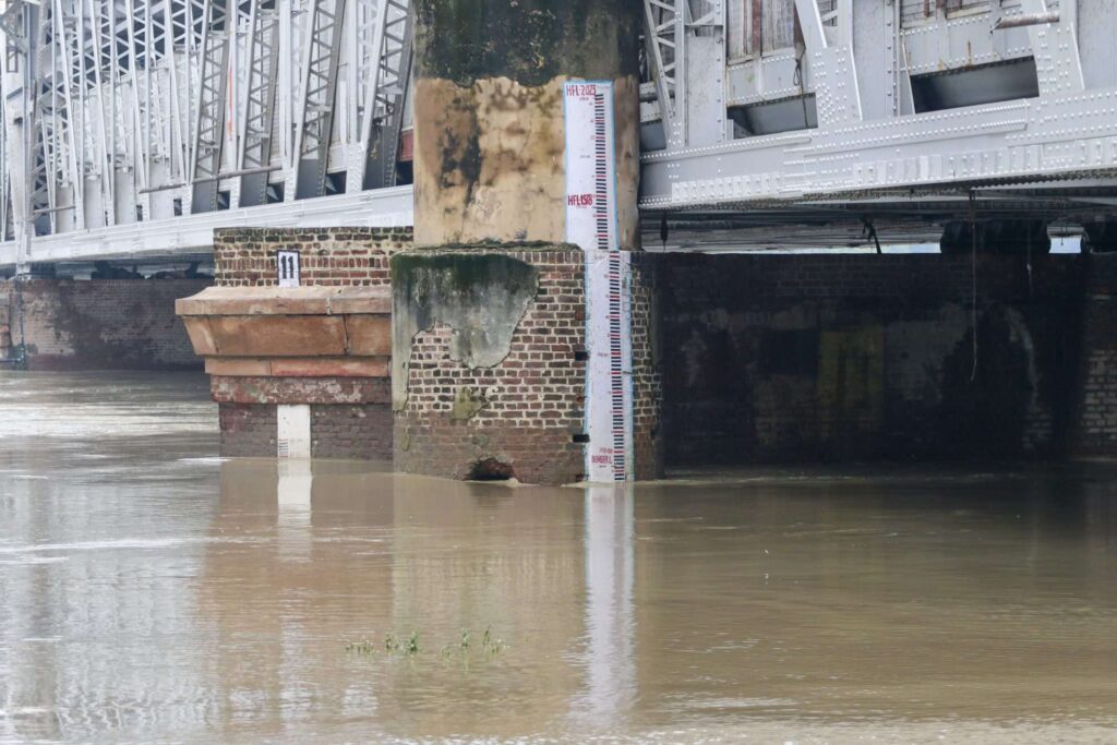 Flood measurement gauge at Old Delhi Railway Bridge showing Yamuna River water level at approximately 207 meters, well above the danger mark, with brownish floodwaters surrounding the brick foundation of the bridge structure.