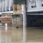 Flood measurement gauge at Old Delhi Railway Bridge showing Yamuna River water level at approximately 207 meters, well above the danger mark, with brownish floodwaters surrounding the brick foundation of the bridge structure.