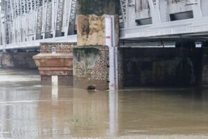 Flood measurement gauge at Old Delhi Railway Bridge showing Yamuna River water level at approximately 207 meters, well above the danger mark, with brownish floodwaters surrounding the brick foundation of the bridge structure.