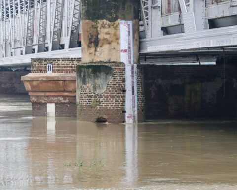 Flood measurement gauge at Old Delhi Railway Bridge showing Yamuna River water level at approximately 207 meters, well above the danger mark, with brownish floodwaters surrounding the brick foundation of the bridge structure.