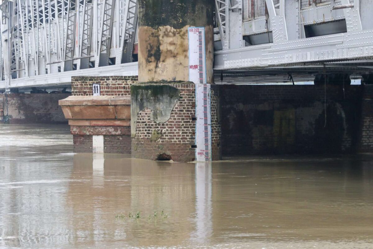 Flood measurement gauge at Old Delhi Railway Bridge showing Yamuna River water level at approximately 207 meters, well above the danger mark, with brownish floodwaters surrounding the brick foundation of the bridge structure.
