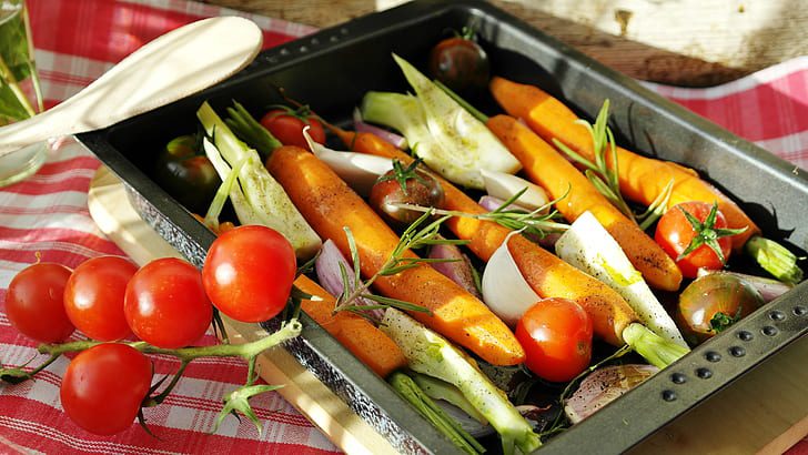 Assorted fresh vegetables including carrots, tomatoes, fennel, and herbs arranged on a stainless steel roasting tray ready for cooking.