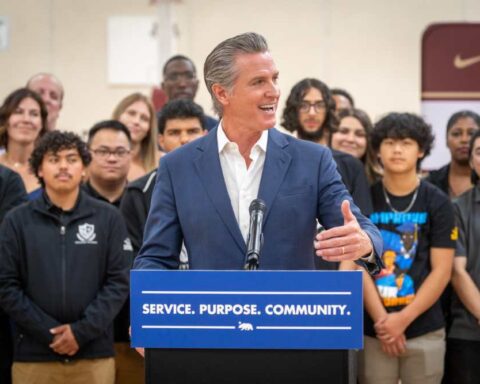 Governor Gavin Newsom speaks at a podium labeled "SERVICE. PURPOSE. COMMUNITY." with young men standing behind him at Florin High School in Sacramento during the launch of the California Men's Service Challenge.