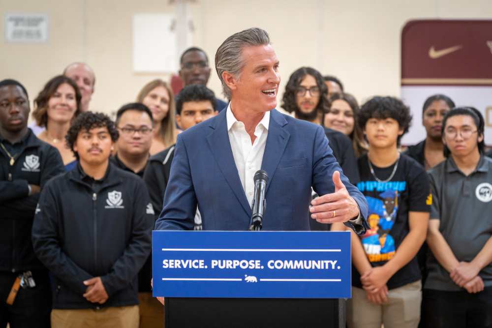 Governor Gavin Newsom speaks at a podium labeled "SERVICE. PURPOSE. COMMUNITY." with young men standing behind him at Florin High School in Sacramento during the launch of the California Men's Service Challenge.