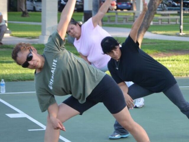 Diverse group of people practicing yoga or stretching exercises outdoors in a park, with five individuals in various side bend poses on a sports court.