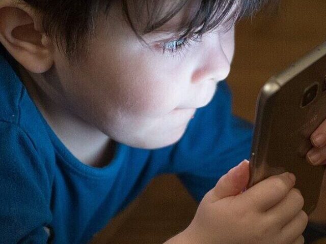 Young boy in blue shirt looking down at smartphone screen while sitting at wooden table