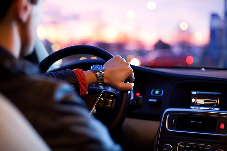 Person driving a car withds on the steering wheel, dashboard and city lights visible in the background