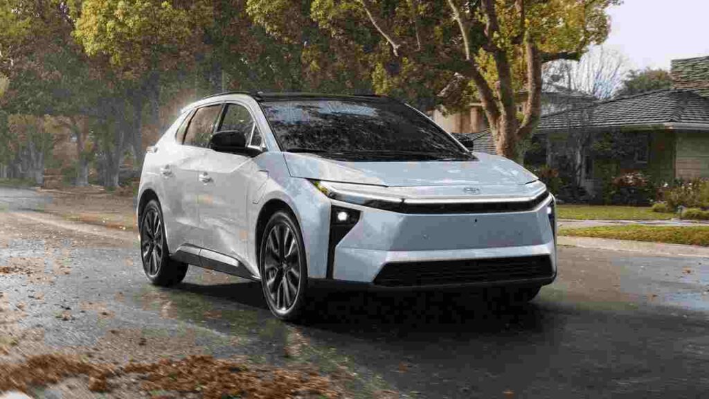 2026 Toyota bZ electric SUV in light blue/silver color driving on a wet residential street with fall leaves, showcasing its modern design with distinctive front lighting and aerodynamic profile.