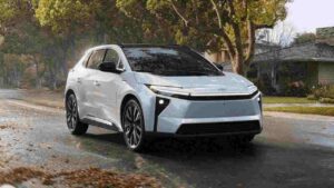 2026 Toyota bZ electric SUV in light blue/silver color driving on a wet residential street with fall leaves, showcasing its modern design with distinctive front lighting and aerodynamic profile.