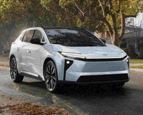 2026 Toyota bZ electric SUV in light blue/silver color driving on a wet residential street with fall leaves, showcasing its modern design with distinctive front lighting and aerodynamic profile.