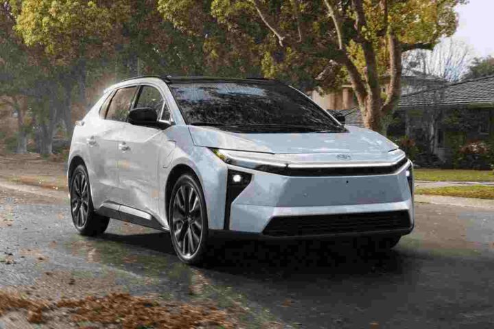 2026 Toyota bZ electric SUV in light blue/silver color driving on a wet residential street with fall leaves, showcasing its modern design with distinctive front lighting and aerodynamic profile.