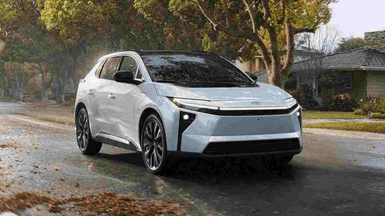 2026 Toyota bZ electric SUV in light blue/silver color driving on a wet residential street with fall leaves, showcasing its modern design with distinctive front lighting and aerodynamic profile.