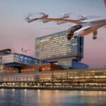 An Archer Midnight eVTOL aircraft flying over the Cleveland Clinic Abu Dhabi hospital at dusk, with the city skyline and water in the background.