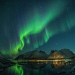 Aurora over a calm lake at night with green arcs reflecting on the water