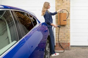 Woman in business attire charging a blue electric vehicle at a wall-mounted home charger