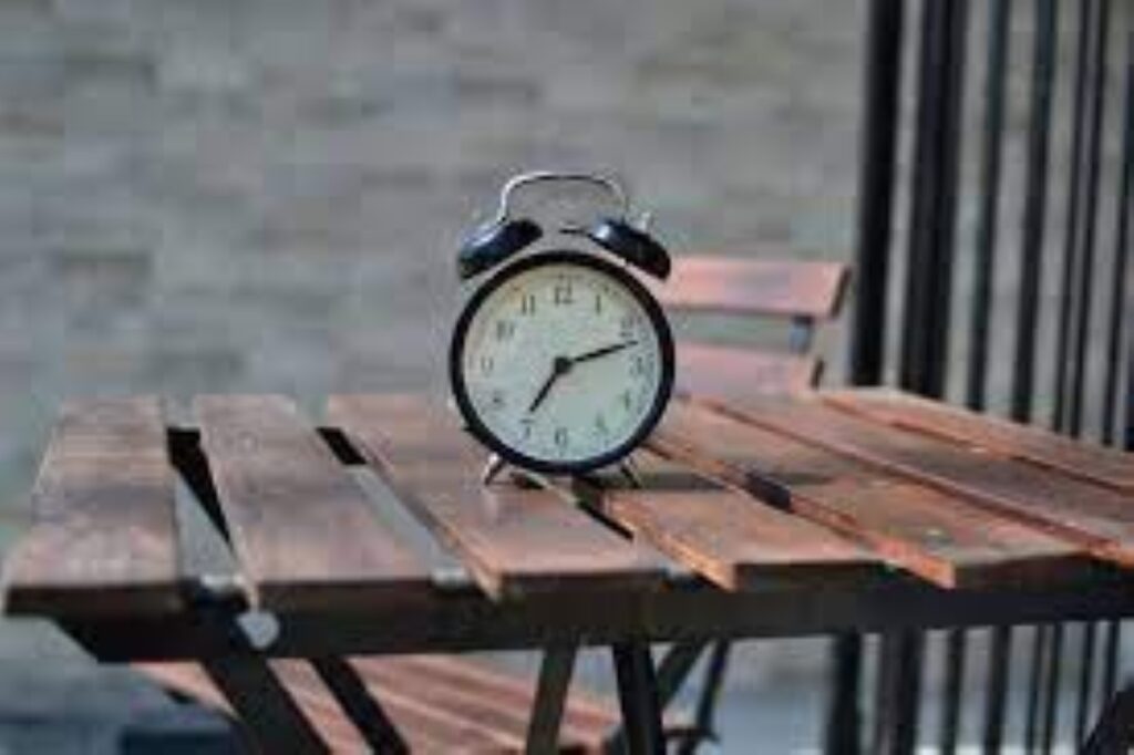 A classic black analog alarm clock sitting on a wooden slatted table with a blurred background.
