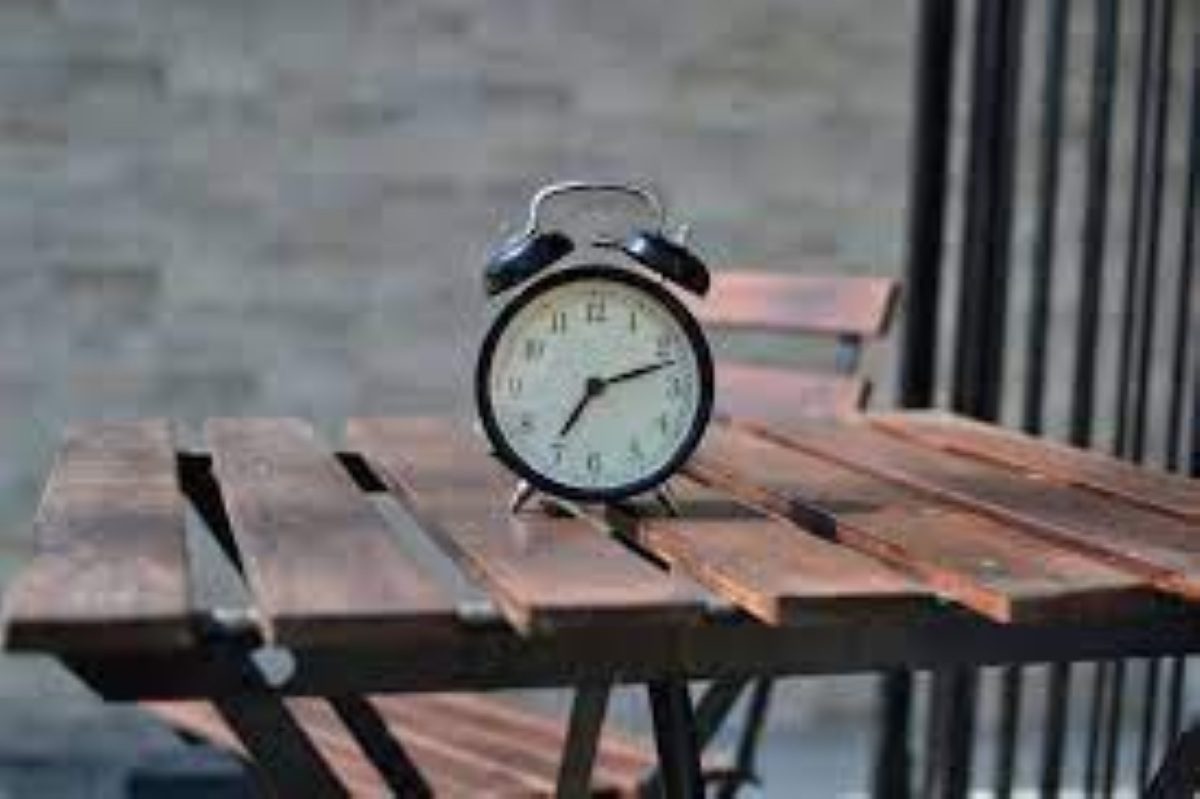 A classic black analog alarm clock sitting on a wooden slatted table with a blurred background.