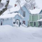 Snow-covered neighborhood street with houses during a winter storm, low clouds, and plowed road.