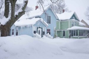 Snow-covered neighborhood street with houses during a winter storm, low clouds, and plowed road.