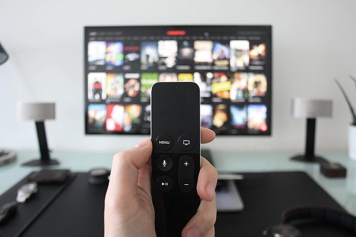 A man sits in a dimly lit room, holding a remote and watching a television screen displaying the Apple TV interface.
