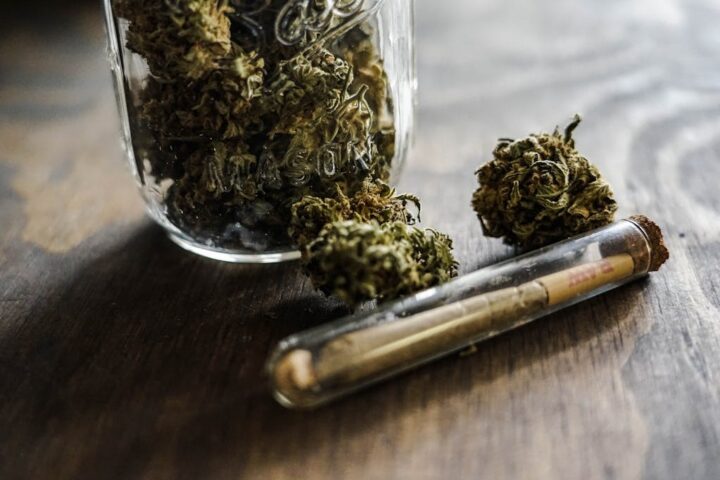 Cannabis buds in a small glass jar with a pre-rolled joint beside it on a wooden surface.