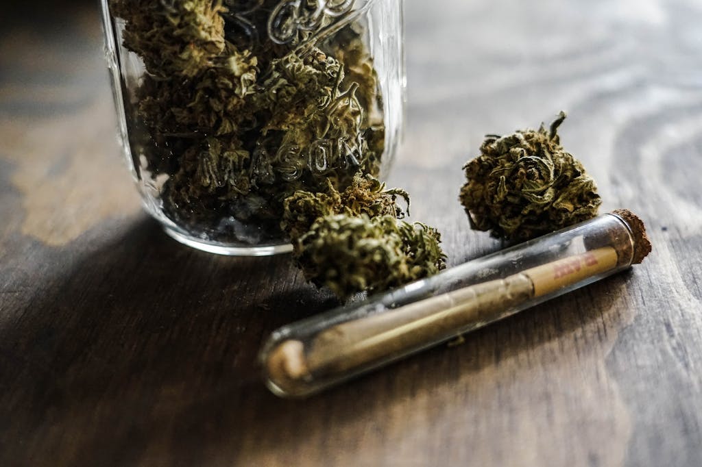 Cannabis buds in a small glass jar with a pre-rolled joint beside it on a wooden surface.