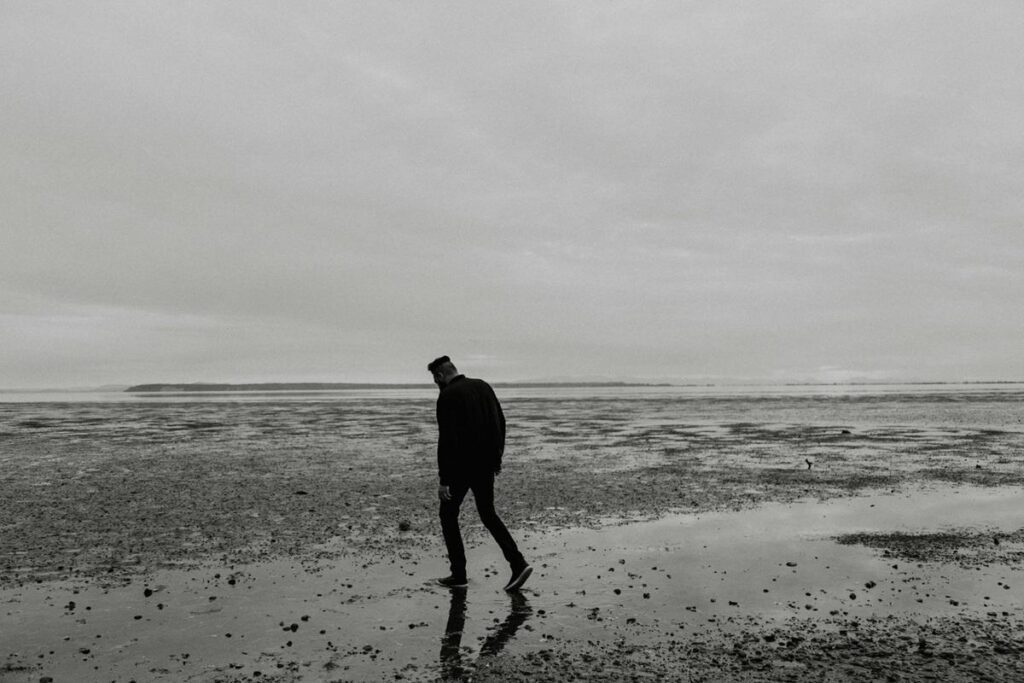 A solitary figure walking alone on a beach shoreline, silhouetted against an overcast sky, representing depression and isolation.