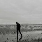 A solitary figure walking alone on a beach shoreline, silhouetted against an overcast sky, representing depression and isolation.