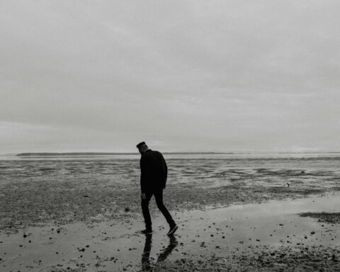 A solitary figure walking alone on a beach shoreline, silhouetted against an overcast sky, representing depression and isolation.