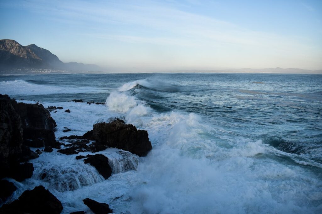 Waves crashing over dark coastal rocks during high tide with distant mountains and a pale blue sky.
