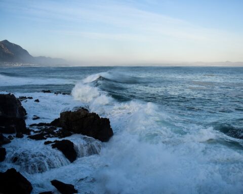 Waves crashing over dark coastal rocks during high tide with distant mountains and a pale blue sky.