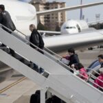 Passengers in a queue at an airport gate walking toward an aircraft jet bridge, carrying luggage and boarding.