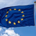 A blue flag with a circle of twelve yellow stars, representing the European Union, waves against a cloudy sky.