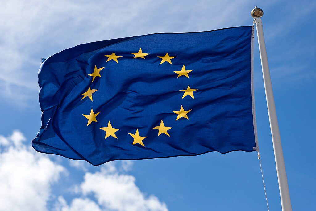 A blue flag with a circle of twelve yellow stars, representing the European Union, waves against a cloudy sky.