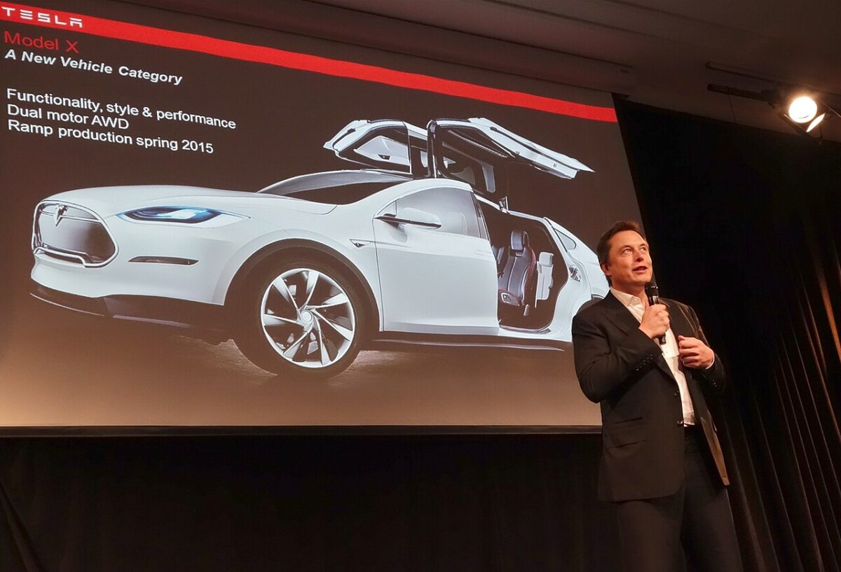 Elon Musk speaking at Tesla's annual shareholder meeting, wearing a black jacket and holding a microphone