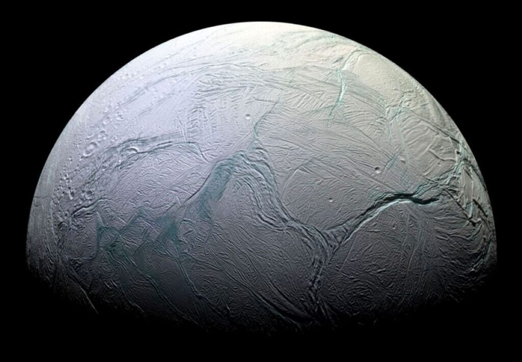 A detailed, close-up image of Enceladus, Saturn's icy moon, showing its heavily cratered and fractured surface with prominent ridges and troughs, illuminated against the darkness of space.