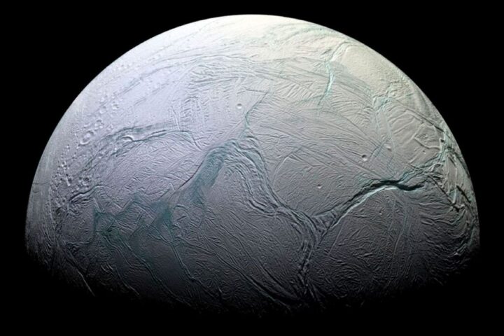 A detailed, close-up image of Enceladus, Saturn's icy moon, showing its heavily cratered and fractured surface with prominent ridges and troughs, illuminated against the darkness of space.