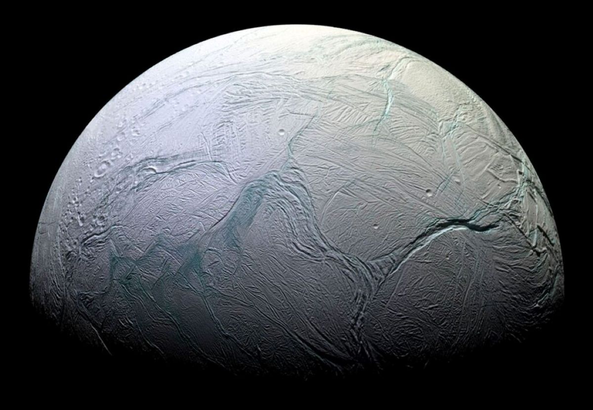 A detailed, close-up image of Enceladus, Saturn's icy moon, showing its heavily cratered and fractured surface with prominent ridges and troughs, illuminated against the darkness of space.