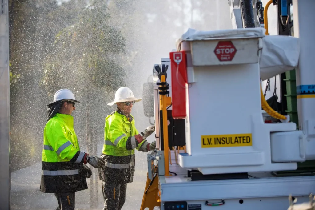 Two Energex lineworkers in hi-vis jackets and hard hats operate equipment next to an LV-insulated service truck while rain sprays around them during post-storm power restoration in South East Queensland.