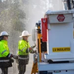 Two Energex lineworkers in hi-vis jackets and hard hats operate equipment next to an LV-insulated service truck while rain sprays around them during post-storm power restoration in South East Queensland.