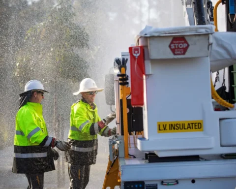 Two Energex lineworkers in hi-vis jackets and hard hats operate equipment next to an LV-insulated service truck while rain sprays around them during post-storm power restoration in South East Queensland.